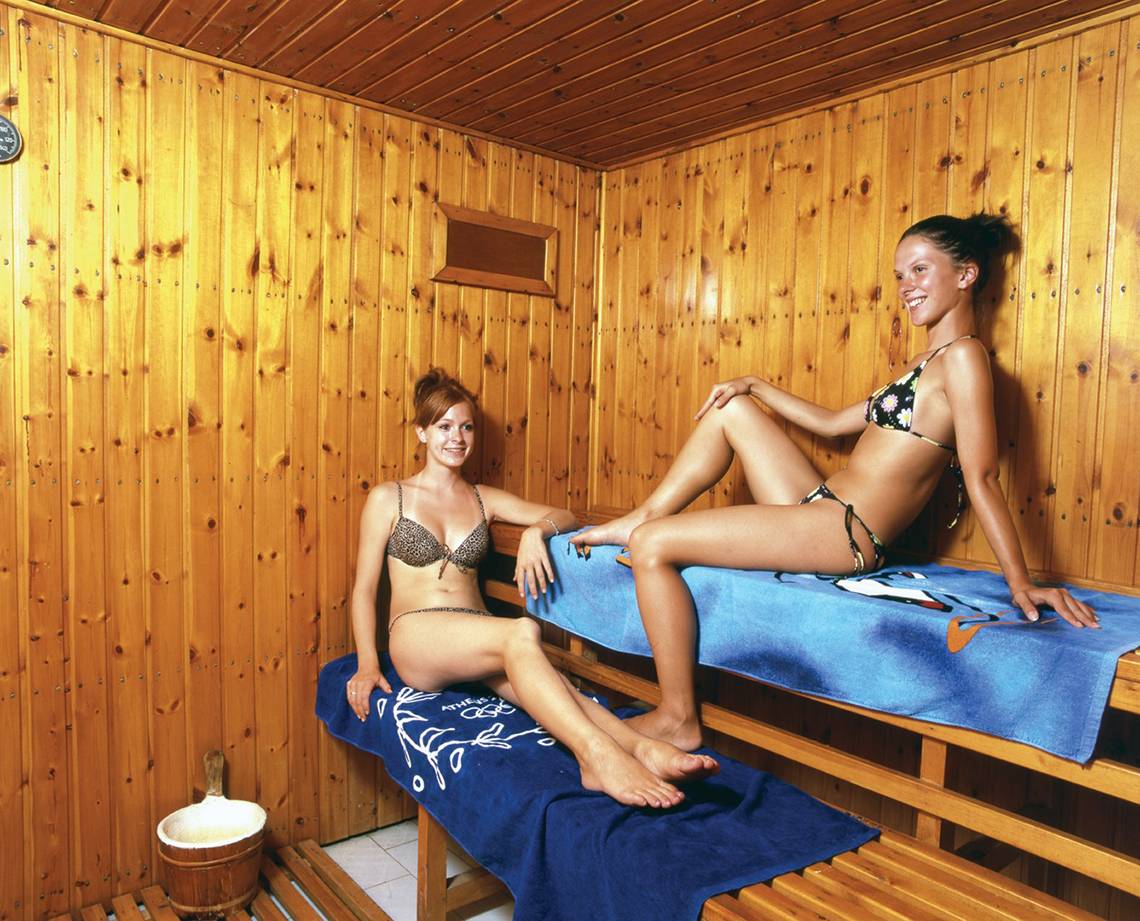 Zwei Frauen in einer Holzsauna, beide tragen Bikinis und lächeln.