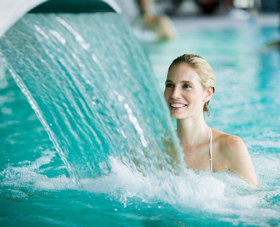 Frau im Schwimmbad unter einem Wasserfall, lächelt ins Bild