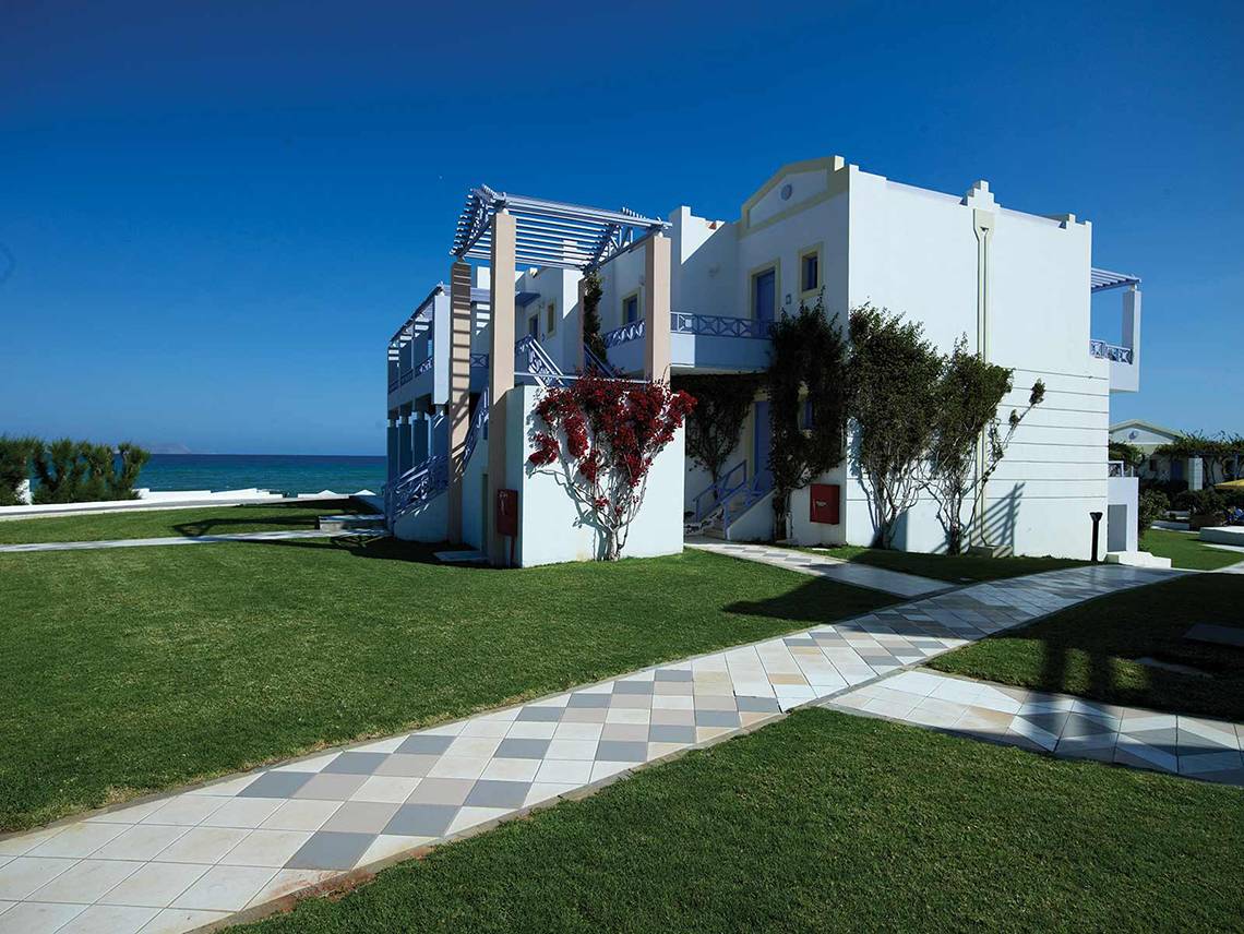 Außenansicht des weißen Serita Beach Hotels mit Rasen, Gehweg und Meer im Hintergrund.