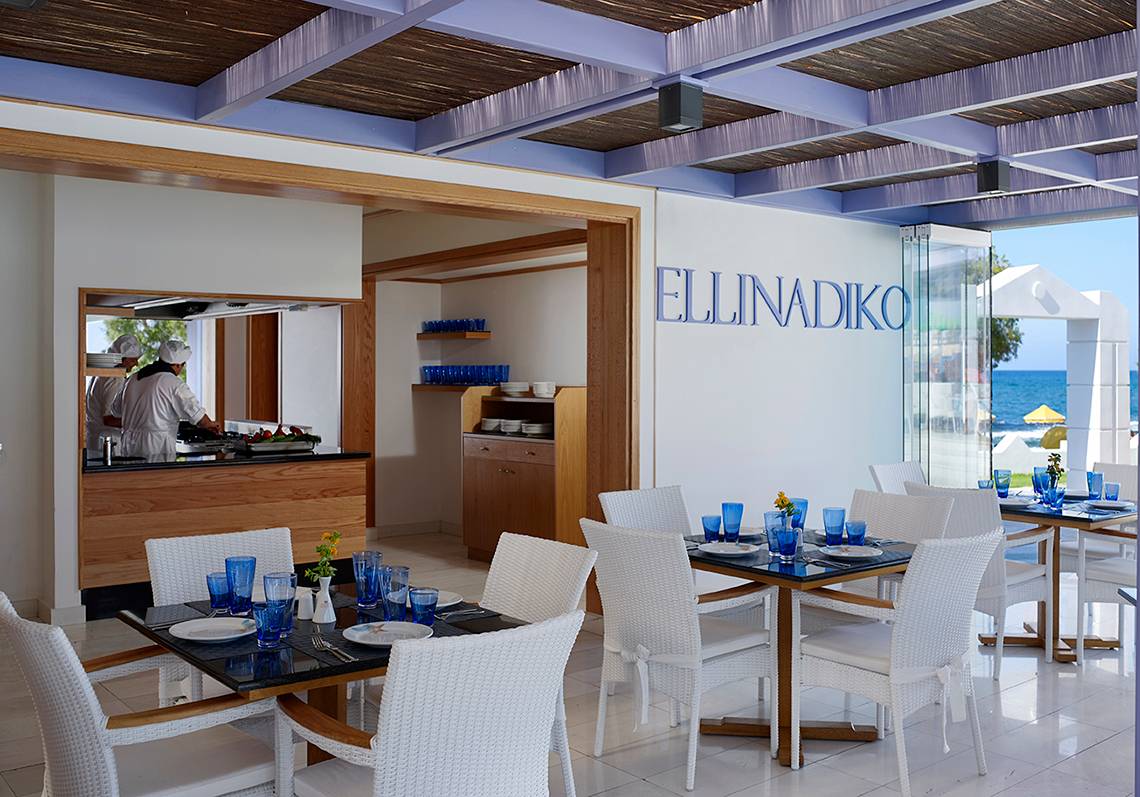 Innenansicht des Ellinadiko Restaurants mit weißen Stühlen und Tischen, Blick auf die Küche und das Meer.