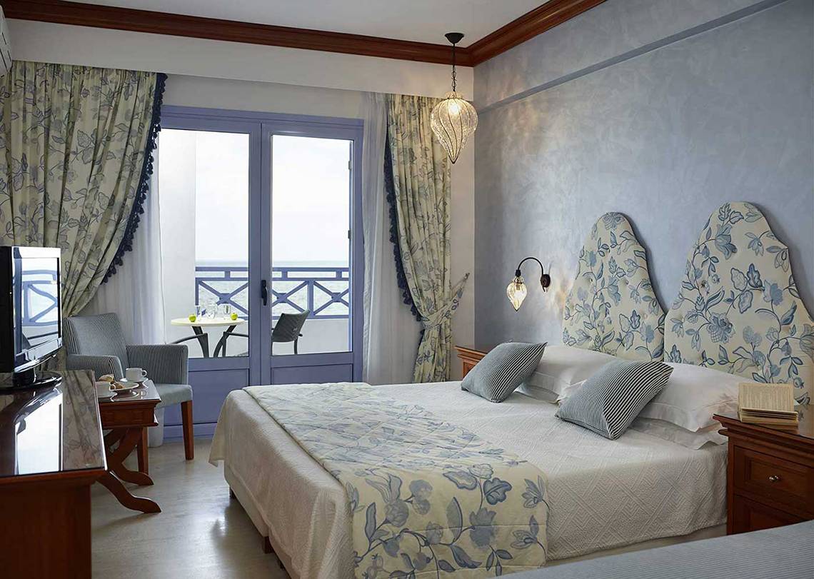 Innenansicht eines luxuriösen Hotelzimmers im Serita Beach Hotel mit einem Doppelbett, Fernseher und Balkon.