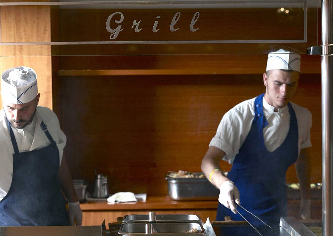Zwei Köche im Grillbereich des Serita Beach Hotels, einer in Uniform, der andere in einer Haube, beide konzentrieren sich auf die Zubereitung von Speisen.