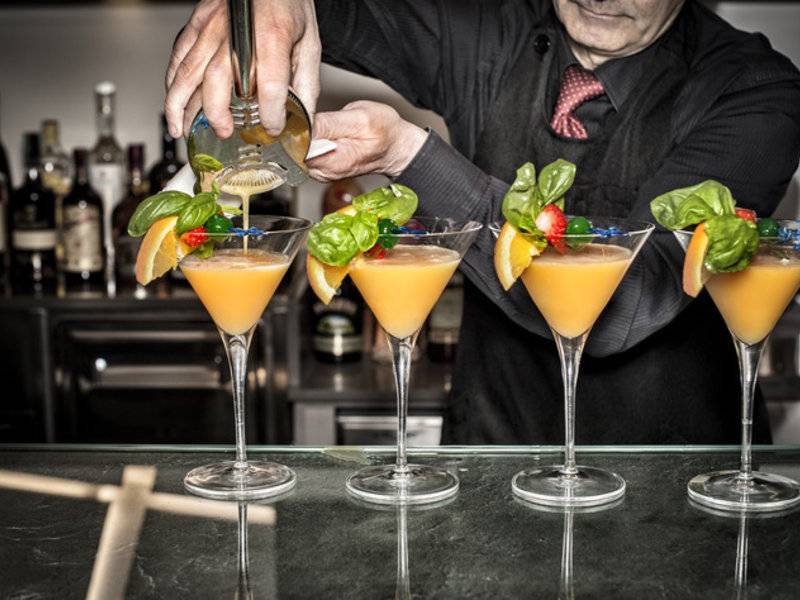 Ein Barkeeper serviert Cocktails auf einer Bar, vier Gläser mit verschiedenen Früchten und Kräutern.