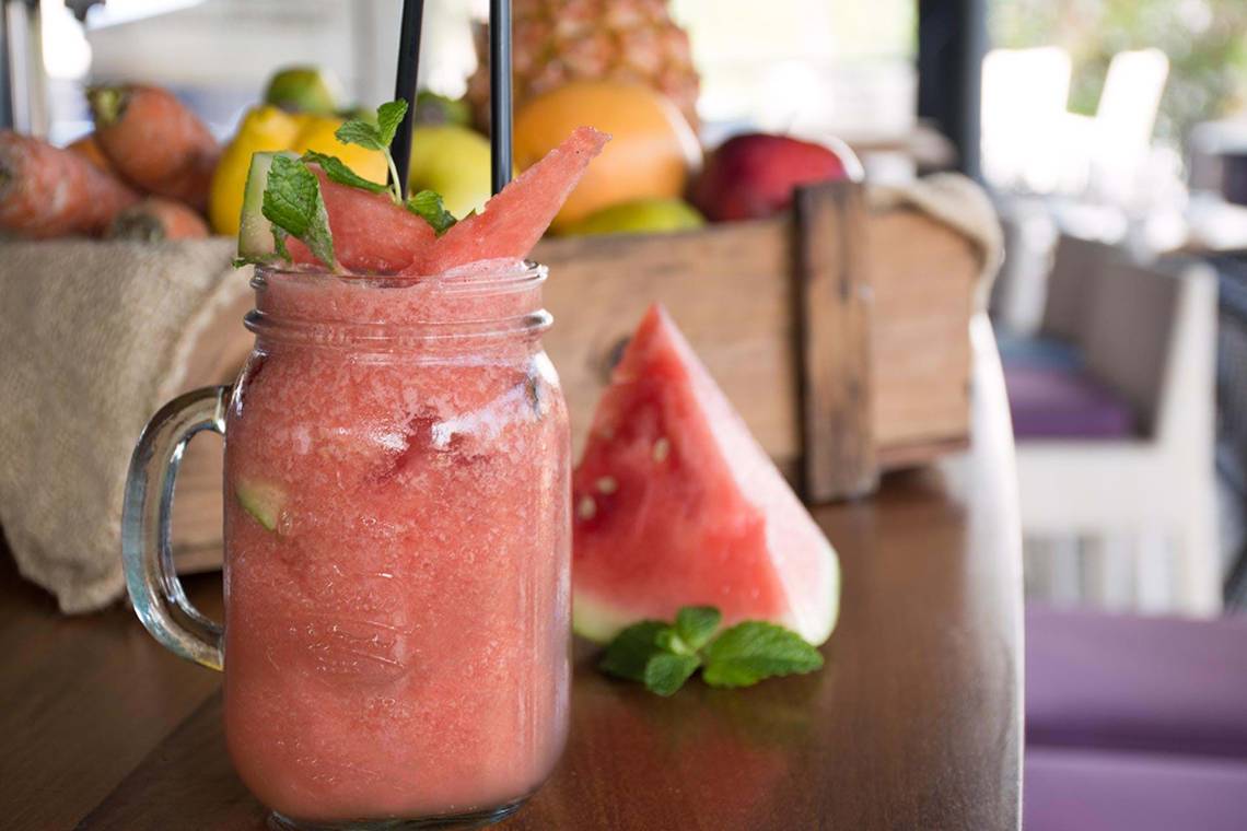 Ein Glas mit einem Wassermelonen-Smoothie auf einem Tisch, mit einem geschnittenen Stück Wassermelone daneben.