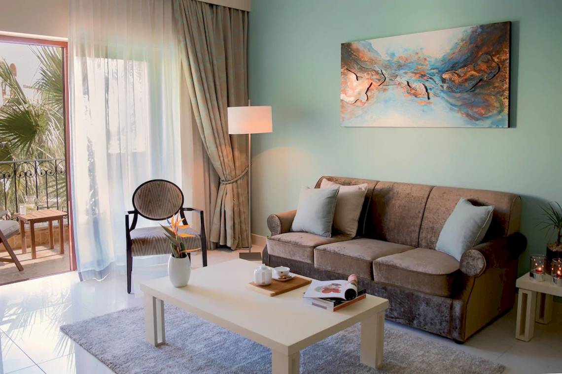 Wohnzimmer im Kempinski Hotel San Lawrenz mit Sofa, Tisch, Gemälde und Balkonaussicht.
