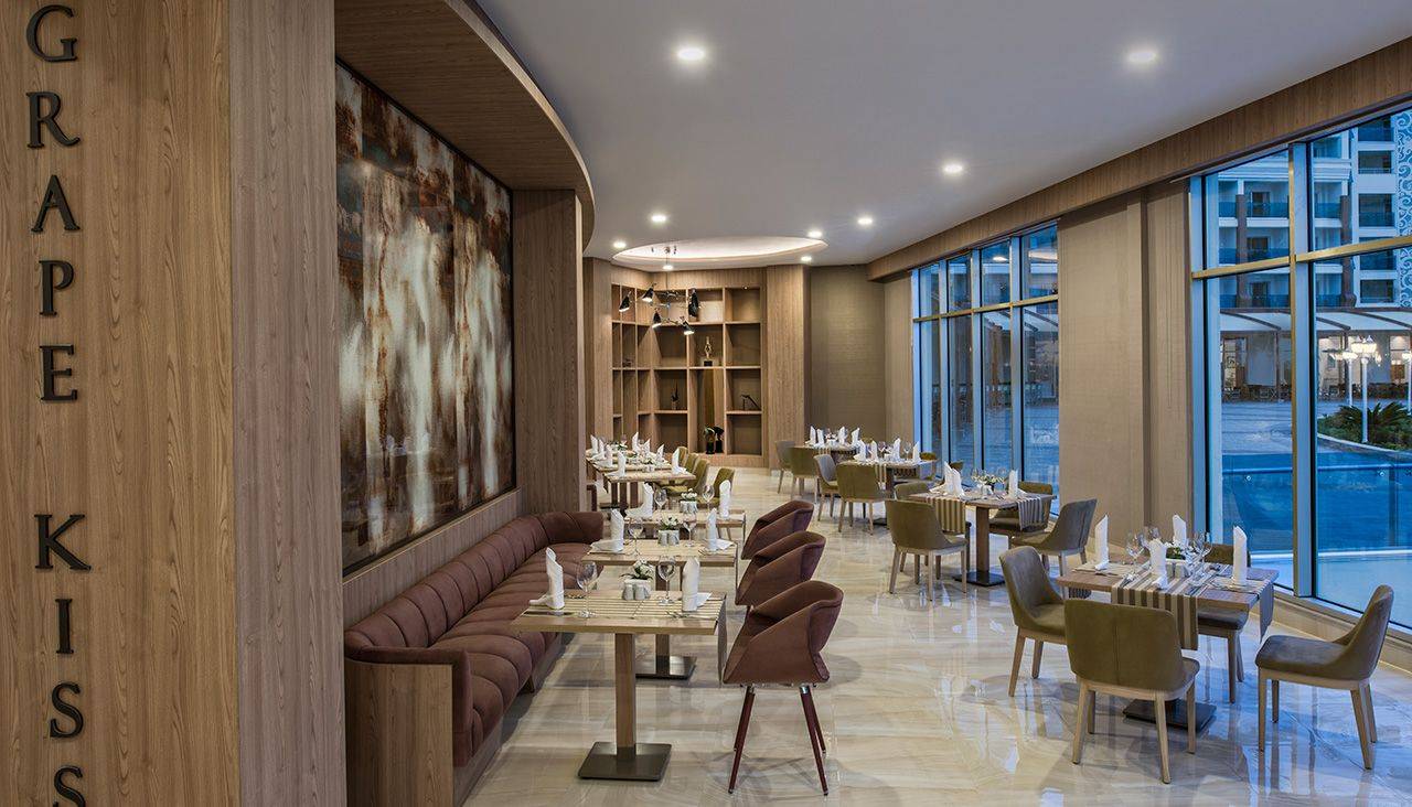 Innenansicht des Grape Kiss Restaurants im Delphin BE Grand Resort mit Tisch und Stühlen, Wandmalerei und großen Fenstern.