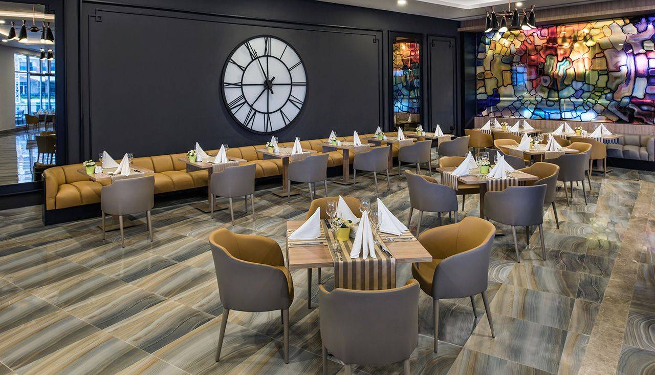 Elegantes Restaurant im Delphin BE Grand Resort mit stilvollen Tischen, bequemen Stühlen und einem großen Wanduhren-Design.