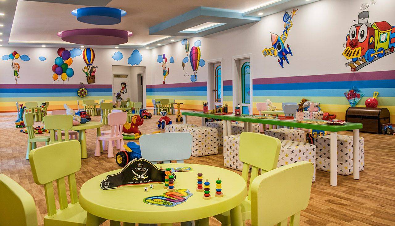 Bunte Spielecke im Delphin BE Grand Resort mit Tischen, Stühlen, Spielzeug und Wandmalereien.