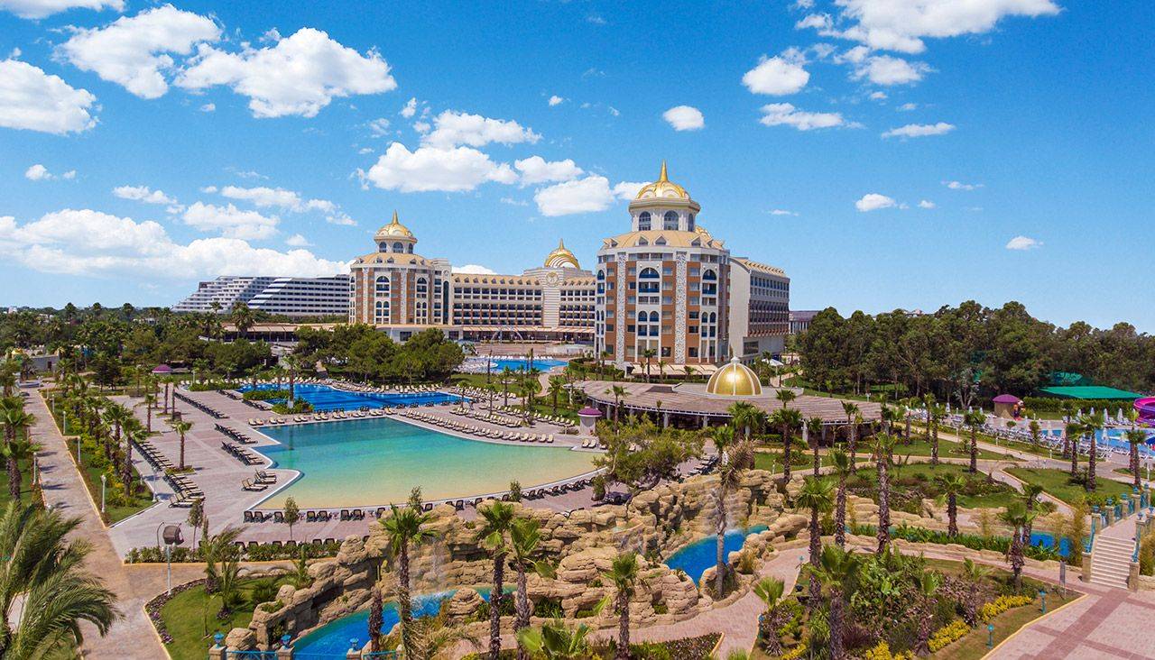 Hotelansicht des Delphin BE Grand Resorts mit Schwimmbad und tropischem Garten unter einem blauen Himmel.