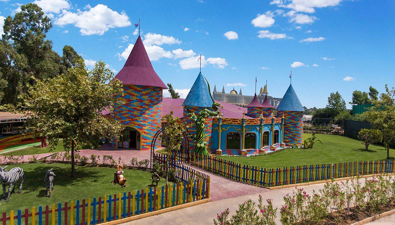 Bunte Architektur des Delphin BE Grand Resort mit Türmen, Garten und Spielplatz, unter blauem Himmel.