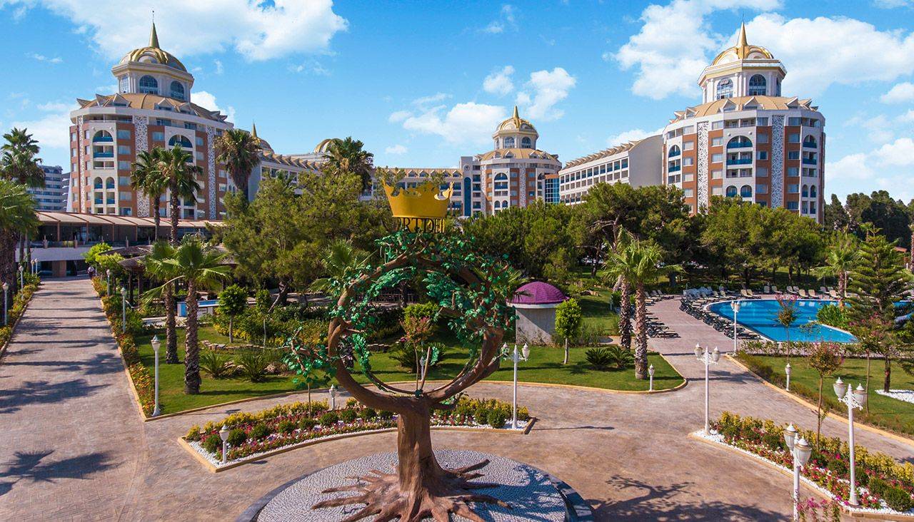 Luxuriöses Delphin BE Grand Resort mit Palmen, Gärten und einer Statue in der Vorderansicht unter klarem Himmel.