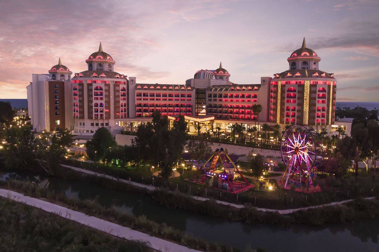 Hotel Delphin BE Grand Resort bei Dämmerung, beleuchtet, mit Riesenrad und Spielplatz im Vordergrund.