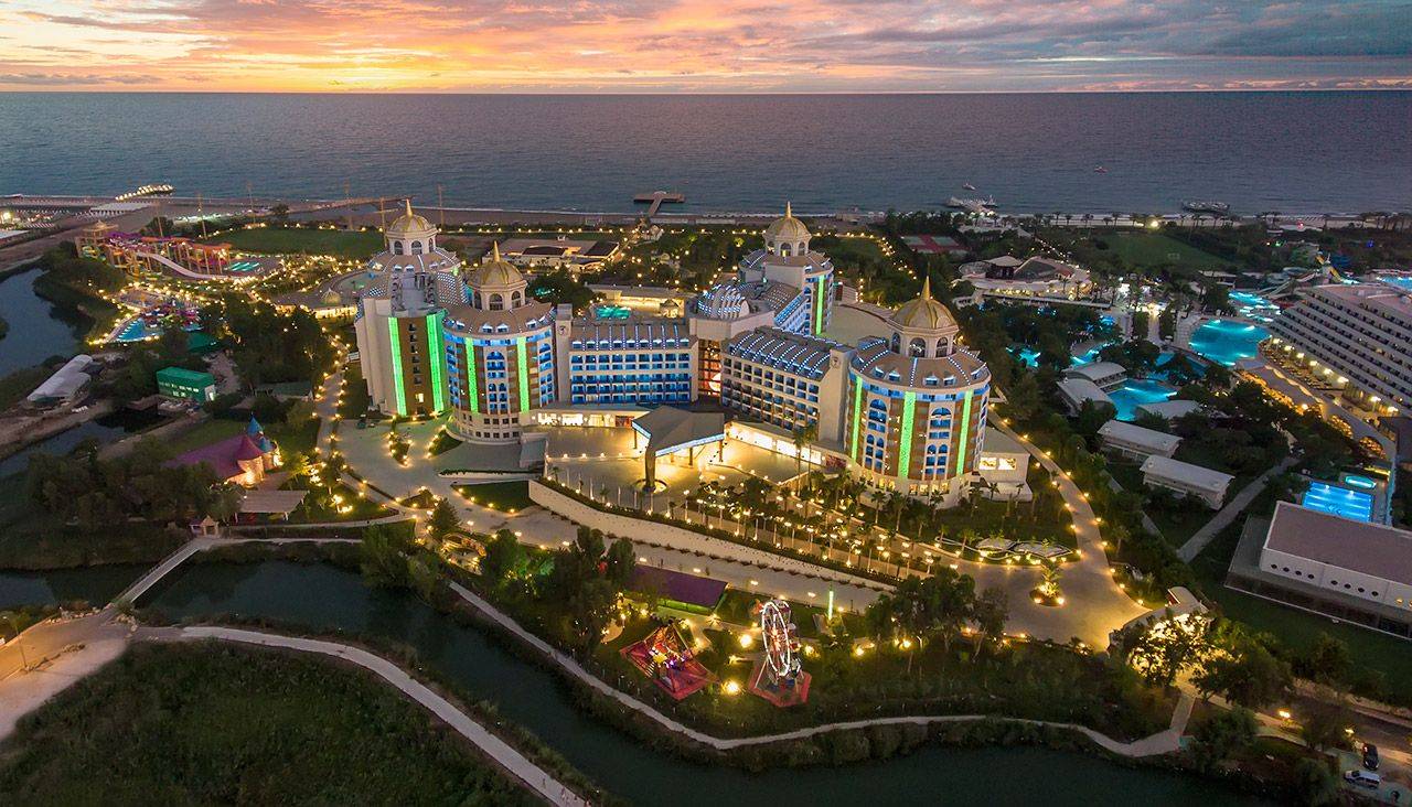 Luftaufnahme des Delphin BE Grand Resort mit bunten Lichtern und Blick auf das Meer bei Sonnenuntergang.