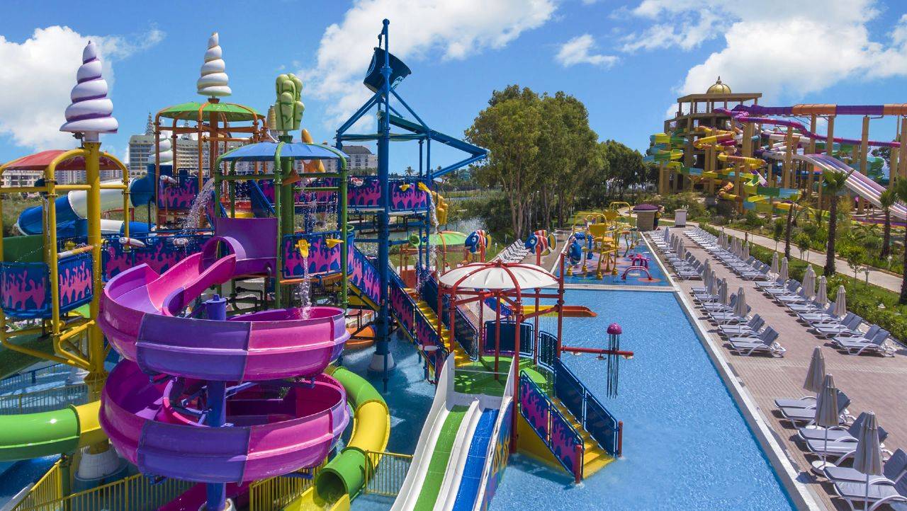 Wasserpark mit bunten Rutschen und Pools im Delphin BE Grand Resort, umgeben von Sonnenliegen und grüner Landschaft.