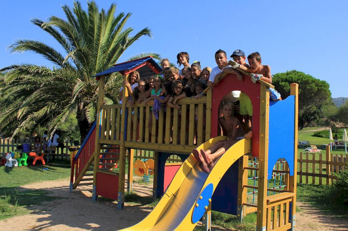 Kinder spielen auf einem bunten Spielplatz im Freien, umgeben von Palmen und einer Holzumzäunung.