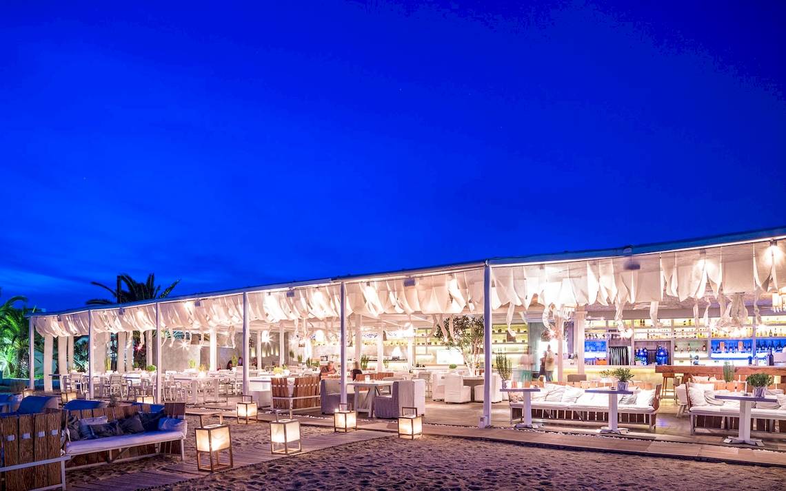 Nachtansicht des Mykonos Ammos Hotel-Strand-Lounge-Bereichs mit weißen Möbeln und Beleuchtung.