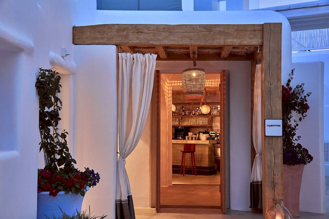 Eingang zum Mykonos Ammos Hotel mit hölzernem Vordach, offenen Türen und Innenansicht des Barsbereichs mit roten Stühlen und Blumentöpfen.