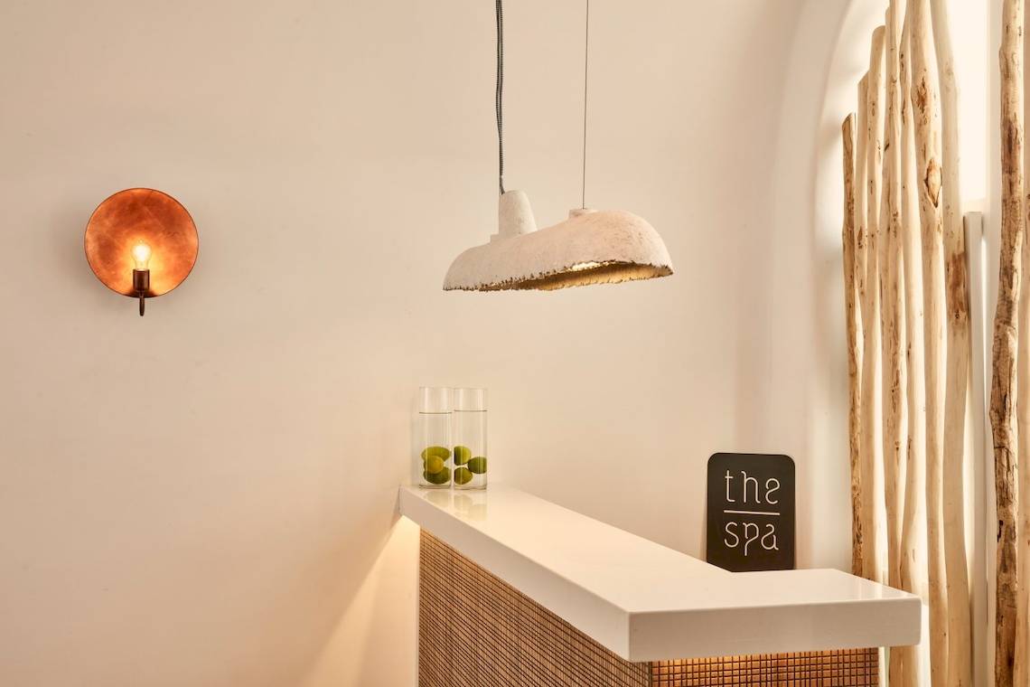 Rezeptionsbereich des Mykonos Ammos Hotels mit einer Wandlampe, einem Glasgefäß mit Früchten und einem Schild mit der Aufschrift 'the spa'.