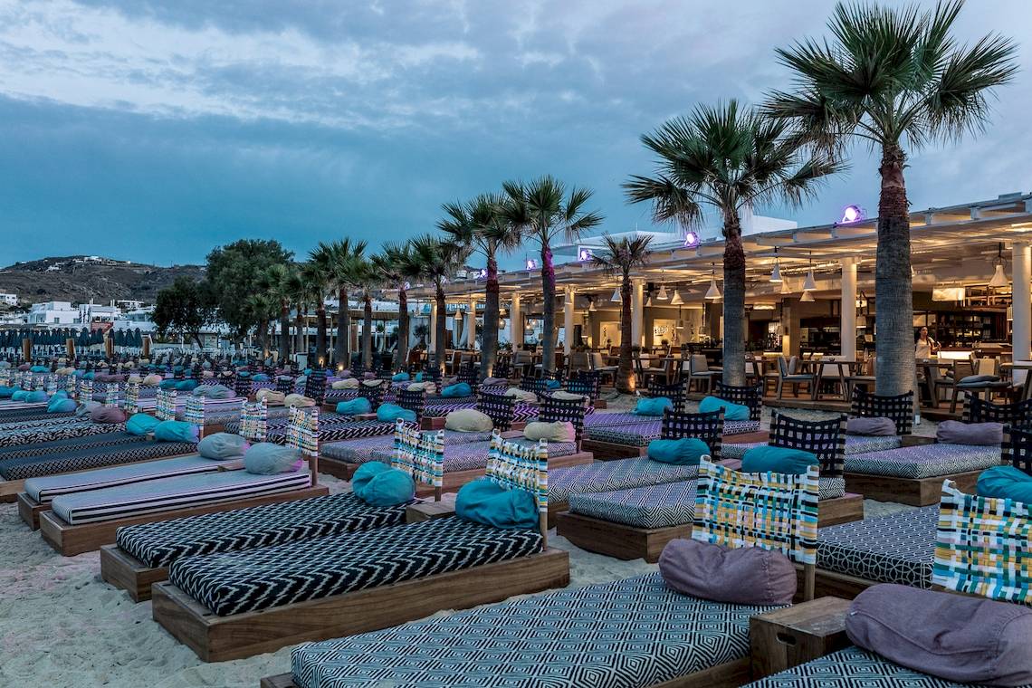 Strandlounge des Mykonos Ammos Hotels mit farbenfrohen Liegen und Palmen.