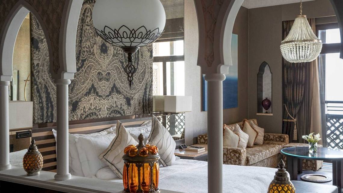 Innenansicht des Schlafzimmers im Jumeirah Mina A'Salam Hotel mit großen Bögen, Kronleuchtern, einem Bett, einem Tisch und einem Sofa.