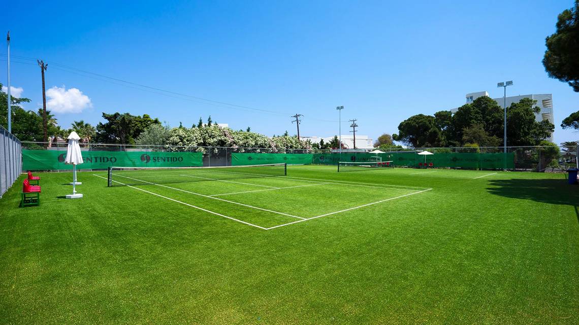 Tennisplatz auf dem Apollo Beach Hotelgelände, mit einem geschlossenen Schirm und grünem Rasen.