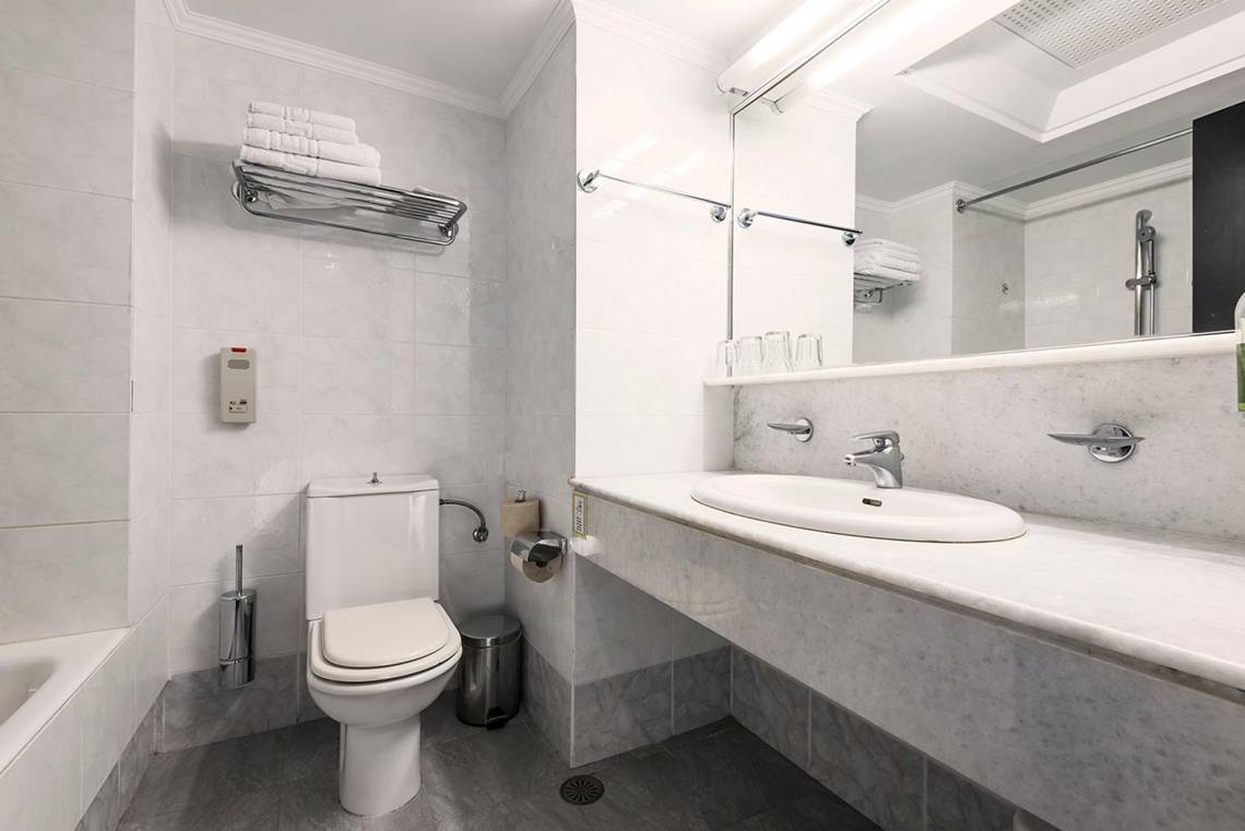 Badezimmer im Apollo Beach Hotel mit weißer Fliesenwand, Toilette, Waschbecken und Spiegel.