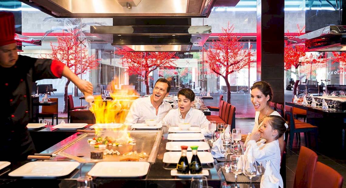 Familie genießt ein Essen in einem Restaurant im Barcelo Maya Grand Resort mit einem Koch, der eine Flamme am Tisch entfacht.