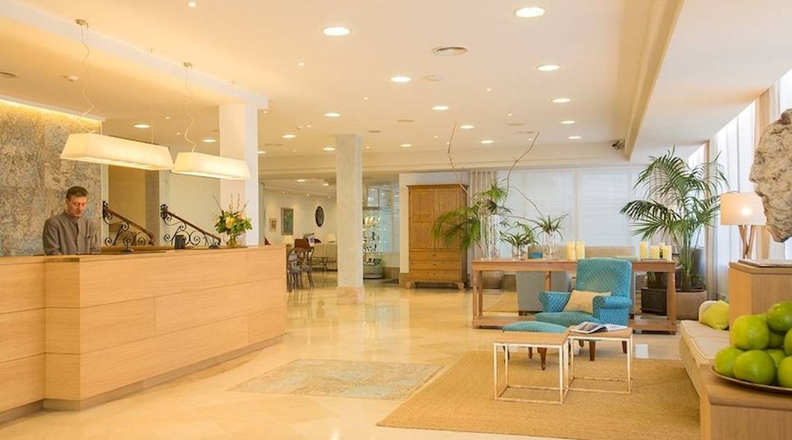 Rezeption im Universal Hotel Neptuno mit hellem Interieur, Holzmöbeln und Pflanzen.