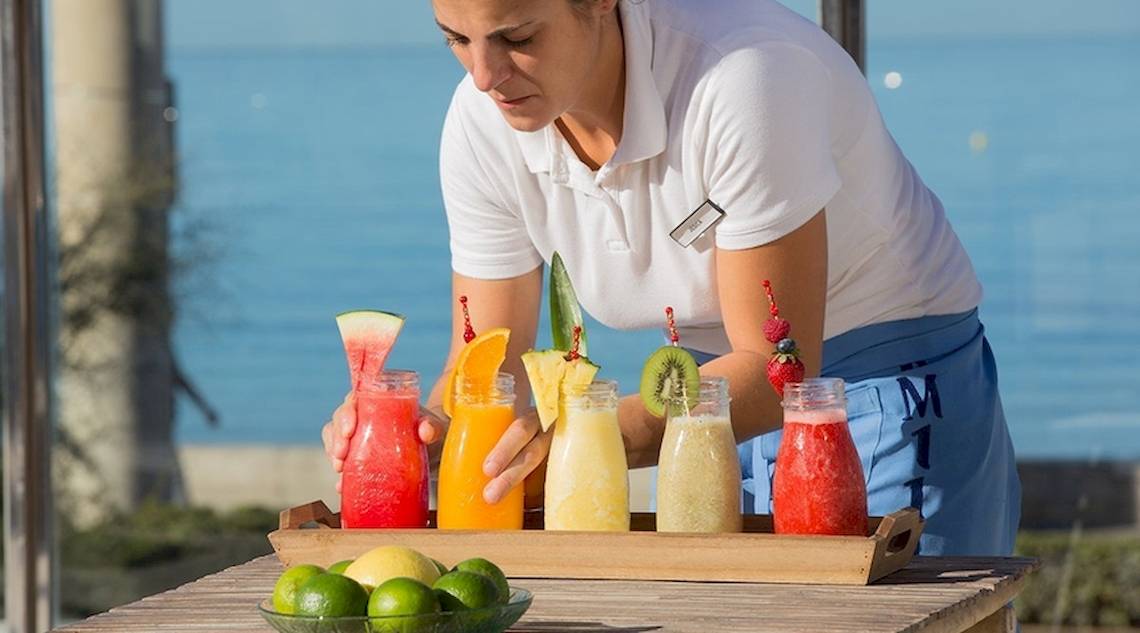 Eine Mitarbeiterin des Universal Hotel Neptuno serviert Cocktails an einem Tisch mit Blick auf das Meer.