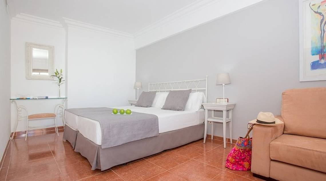 Schlafzimmer im Universal Hotel Neptuno mit weißem Bett, grauen Kissen, grünen Äpfeln, zwei Nachttischen, Tischlampe und einem Spiegel.