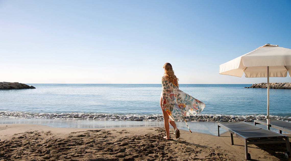 Eine Frau am Strand des Royal Apollonia Beach Hotels mit einem weißen Sonnenschirm im Hintergrund.