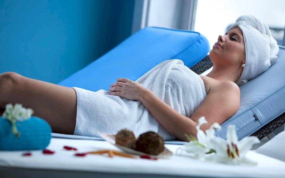 Eine Frau entspannt sich im Spa-Bereich des Seaside Los Jameos, mit einem Handtuch auf dem Kopf und auf einem blauen Liegestuhl.