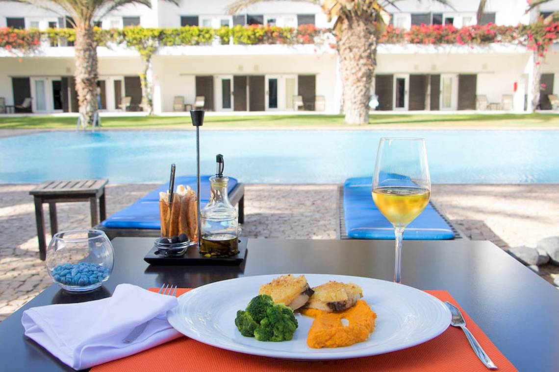 Gedeckter Tisch am Pool mit Essen, Wein und Hotel Dunas De Sal im Hintergrund.