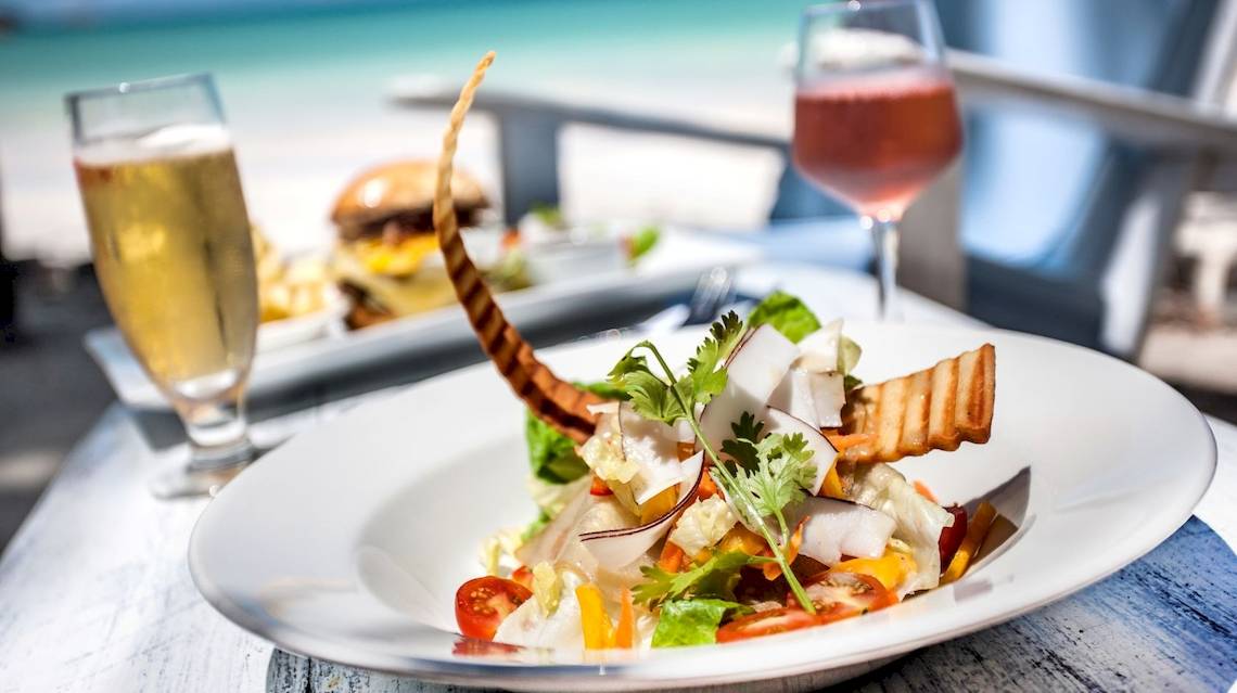 Ein Teller mit Salat und anderen Gerichten auf einem Tisch am Strand mit Getränken und einer schönen Aussicht.