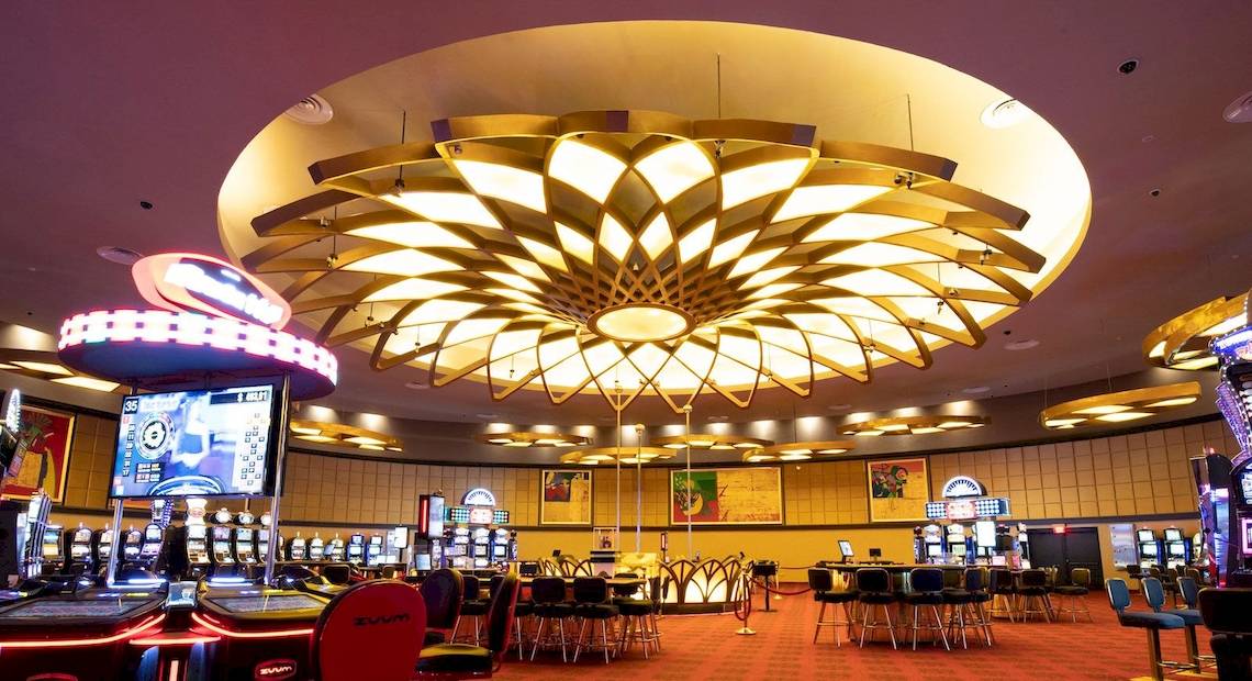 Innenbereich eines Casinos mit Spieltischen, Spielautomaten und einer großen, blumigen Lampe an der Decke.