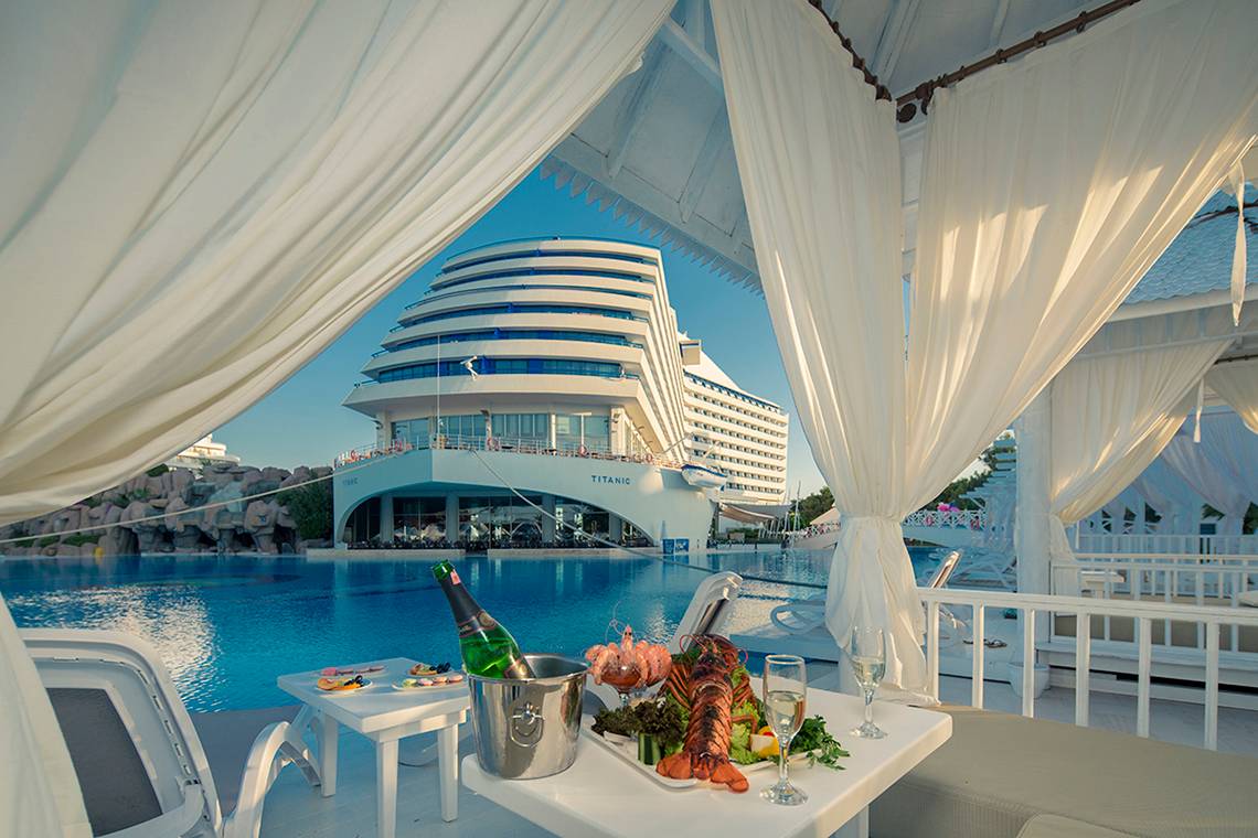 Tisch im Hotelpool-Loungebereich mit Getränken und Essen, Titanic Deluxe Lara im Hintergrund.