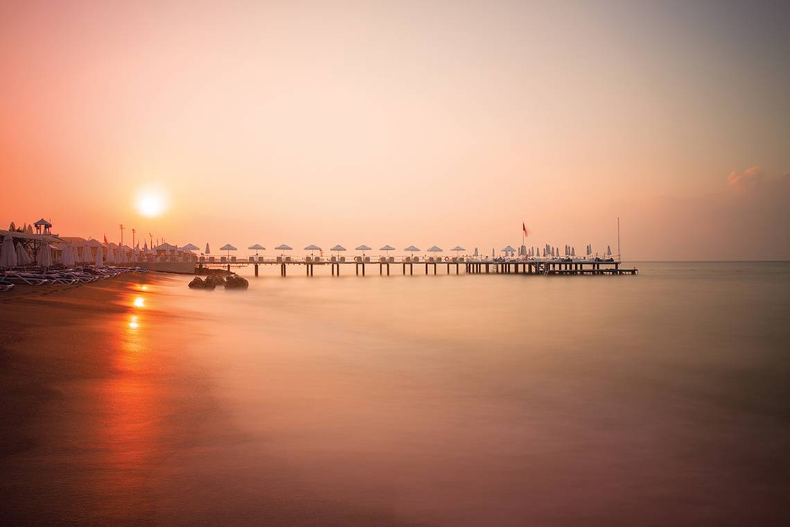 Ein Strand mit einem Pier und dem Sonnenuntergang im Hintergrund, reflektiert auf dem Wasser.