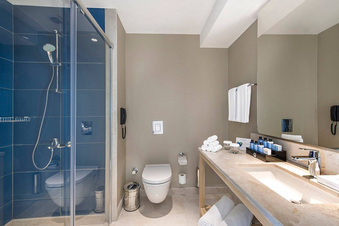 Badezimmer des Titanic Deluxe Lara Hotels mit blauen Kacheln, Dusche, Toilette und Waschbecken.
