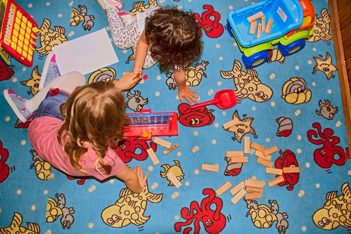 Zwei Kinder spielen mit Holzsteinen auf einem bedruckten Teppich.