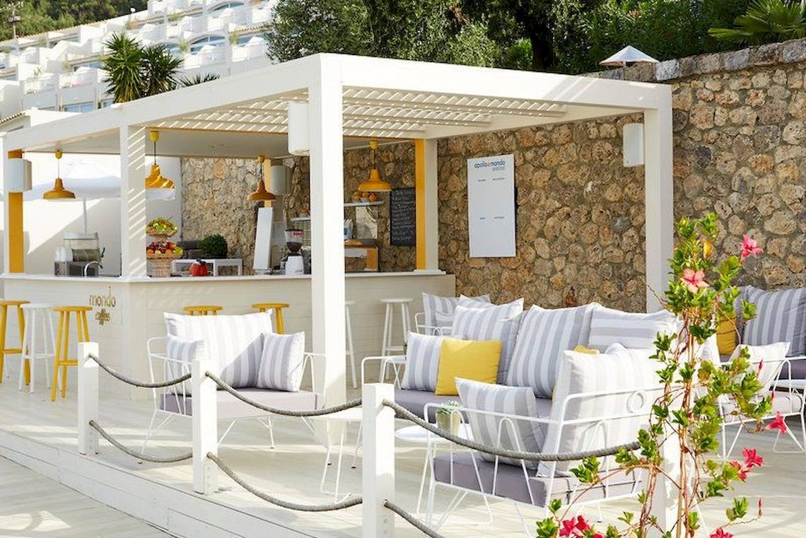 Außenbereich des Pelekas Monastery Hotels mit einer weißen Pergola, gelben Stühlen und Tischen.