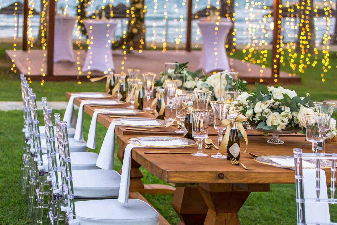 Ein Banketttisch im Freien beim Golden Bay Beach Hotel mit dekorierten Tischen und Stühlen, Gläsern, Kerzen und Blumen.