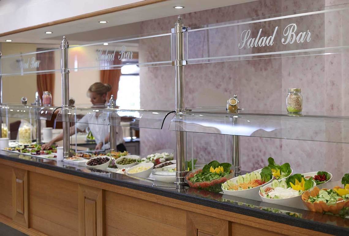 Salatbar im Mitsis Rinela Beach Hotel mit verschiedenen Salaten und einer Mitarbeiterin.