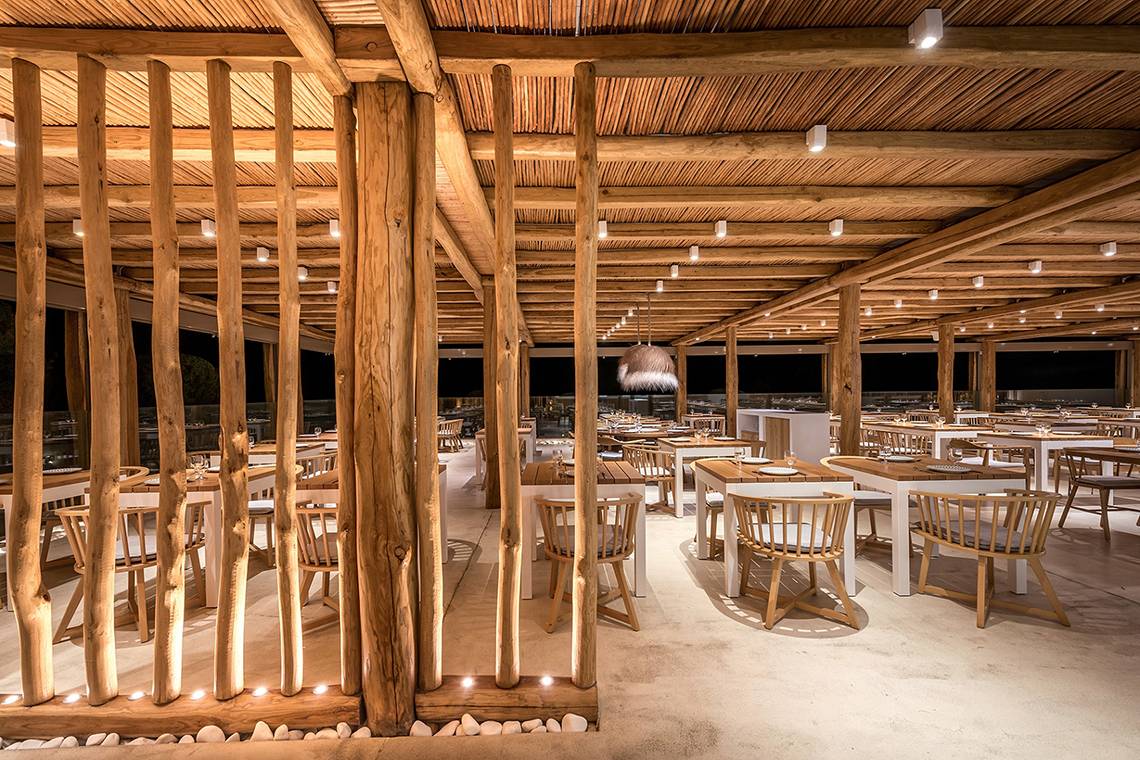 Innenraum der Terrasse des Mitsis Rinela Beach Hotels mit hölzernen Säulen und Tischen.