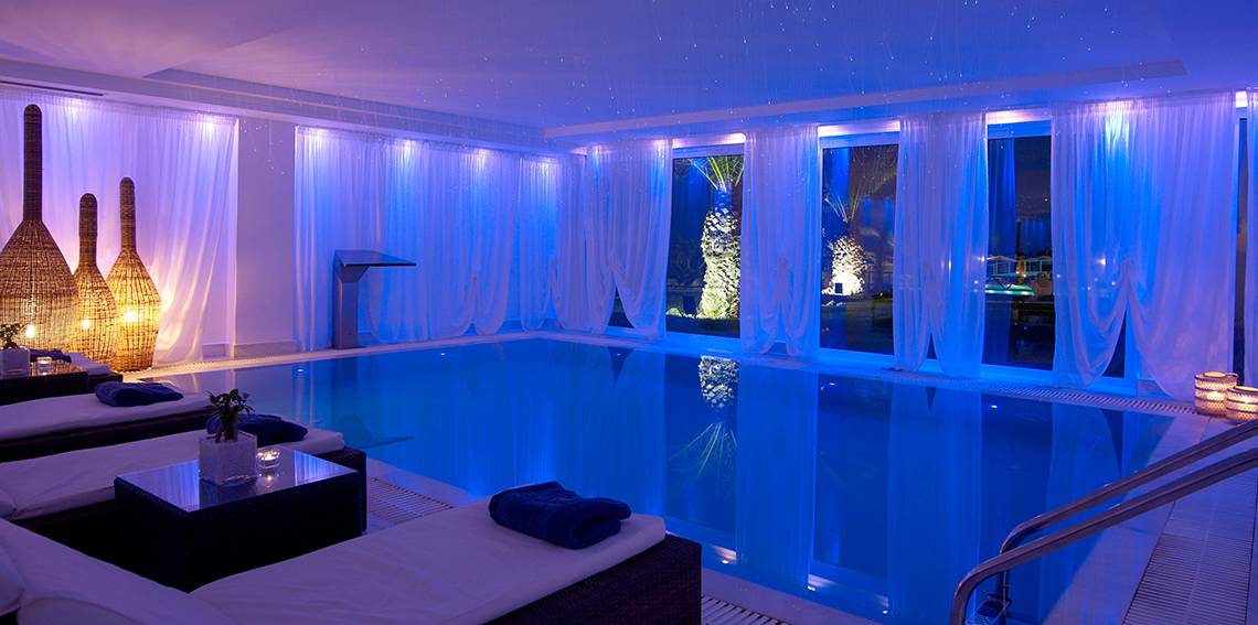 Poolbereich des Mitsis Rinela Beach Hotels mit blauer Beleuchtung und weißen Vorhängen.