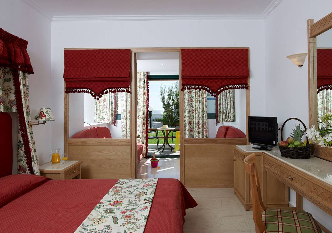 Innenansicht eines Zimmers im Mitsis Rinela Beach Hotel mit roten Vorhängen, einem Bett und einem Schreibtisch.