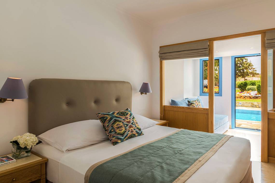 Innenansicht eines Schlafzimmers im Mitsis Rinela Beach Hotel mit Bett, Dekor und offenem Balkon.
