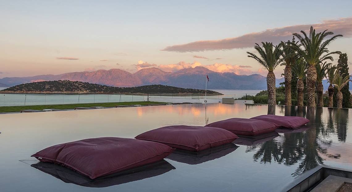 Minos Palace Hotel & Suites: Ein Swimmingpool mit roten Kissen und Blick auf eine bergige Landschaft und einen Sonnenuntergang.