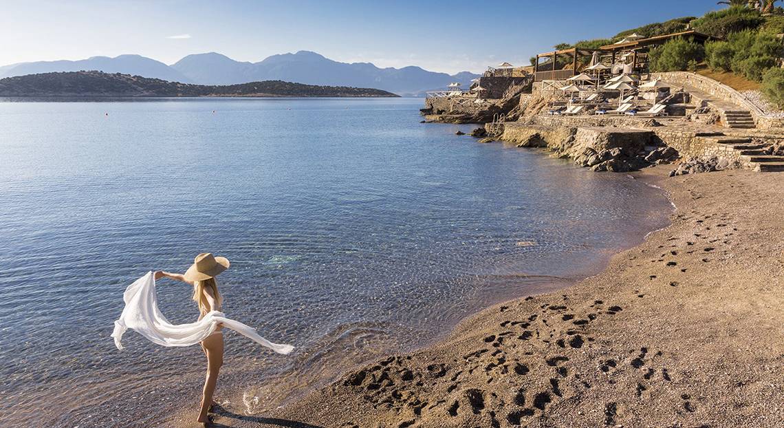 Eine Frau in einem Bikini und Hut steht am Strand des Minos Palace Hotel & Suites.