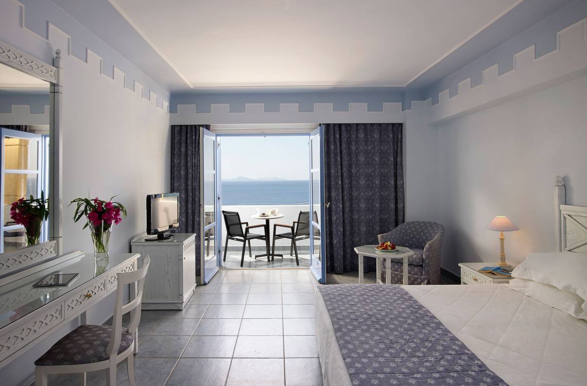 Zimmer im Mitsis Summer Palace Beach Hotel mit Meerblick, Bett, Schreibtisch, Stuhl, Fernseher und Balkon mit Tisch und Stühlen.