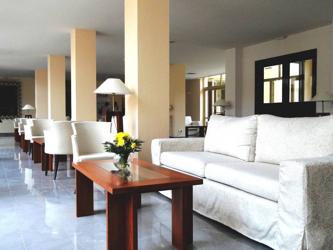 Hotellobby mit Sofa, Tisch mit Blumen, weißen Sesseln und Säulen im Hintergrund.